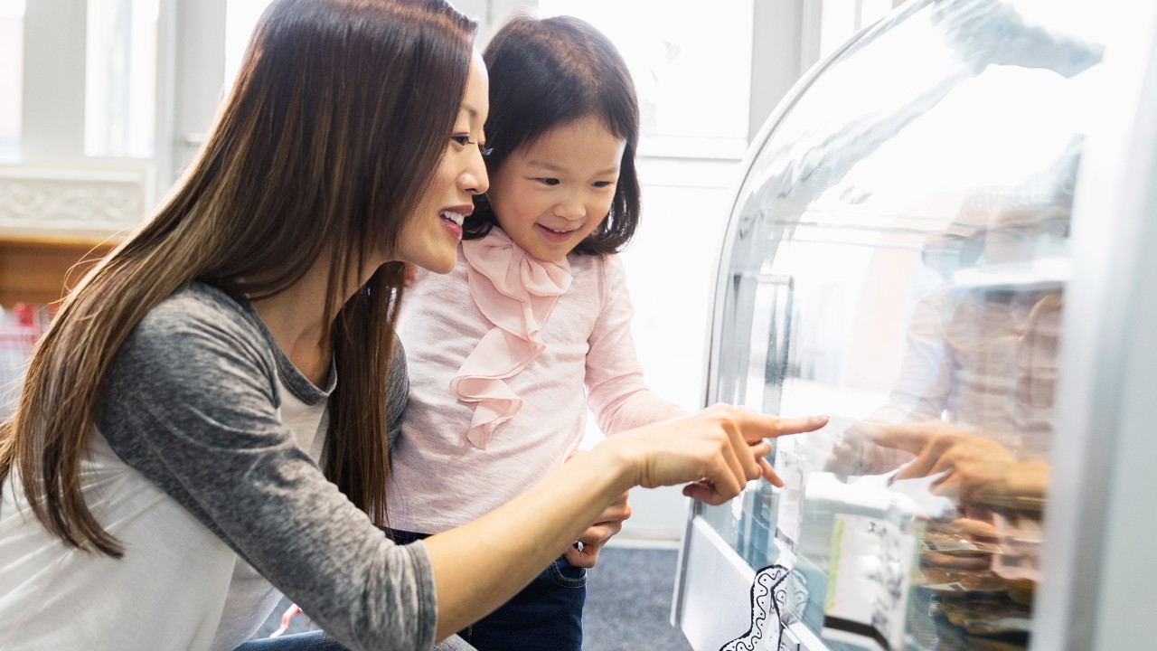 a mother holding her daughter and pointing to the food in a glass display cabinet; image used for HSBC visa gold card page.