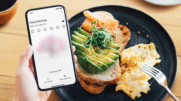 bove a dining table with a hearty breakfast, a hand holding a mobile phone with the HSBC mobile banking app open; image used for HSBC Macau Mobile Banking page